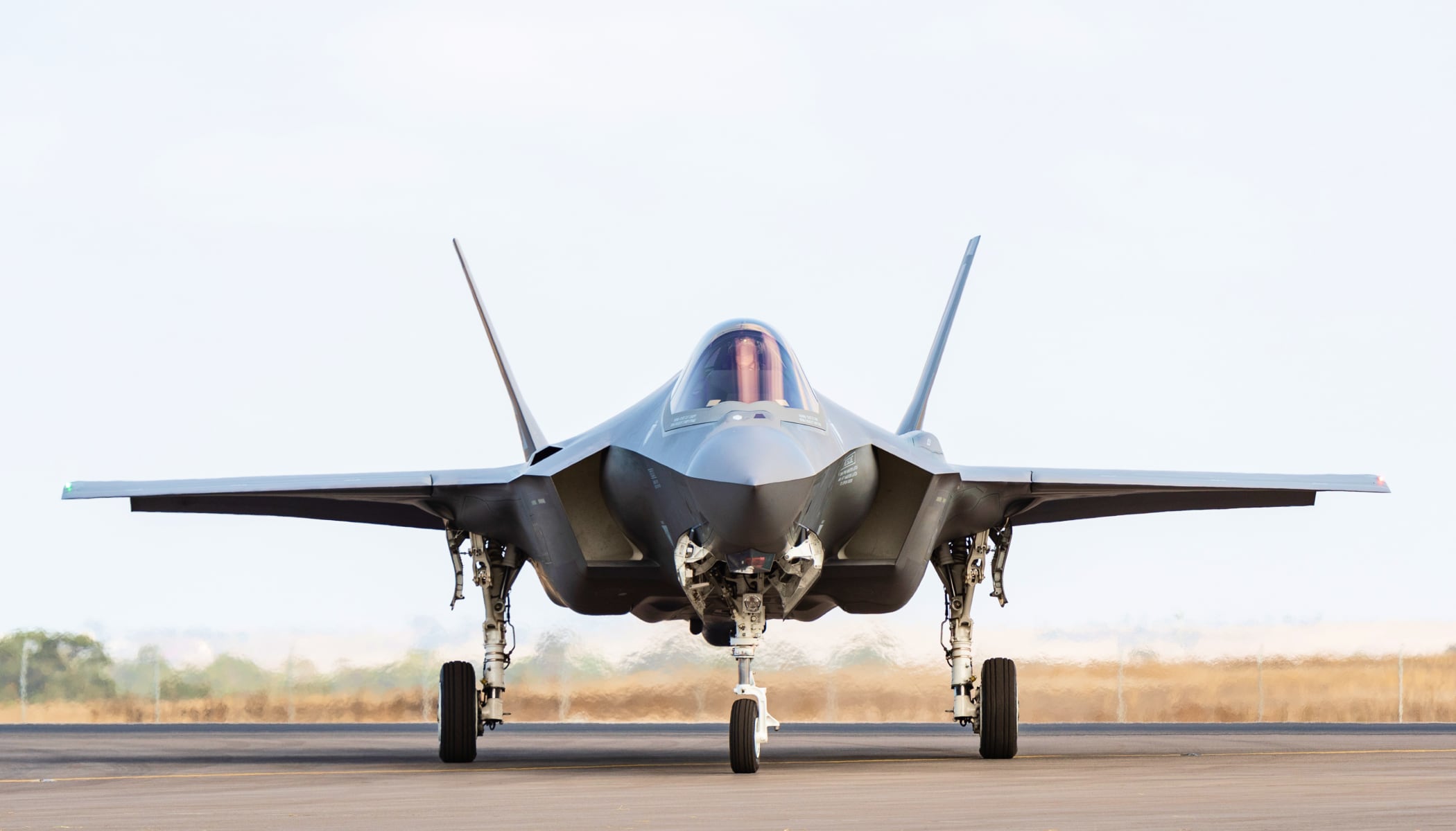 A photo of a modern gray fighter jet parked on a runway ready for flight, taken from the front with the nose of the craft pointed at the viewer. A flat brown field under a pale sky is visible in the background